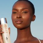 Deep-tone brunette model with glowy, luminous skin, having applied a glowy toner and holding up a bottle of it.