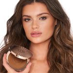 Medium-tone brunette model with brown eyes wearing nude pink lip gloss with glowy, cream bronzed for a sculpted yet natural makeup look while holding up an open bronzer compact.