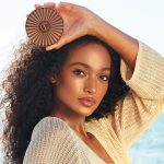 Deep-tone brunette model with glowy bronzed skin and soft-glam pink makeup holding a closed cream bronzer compact.