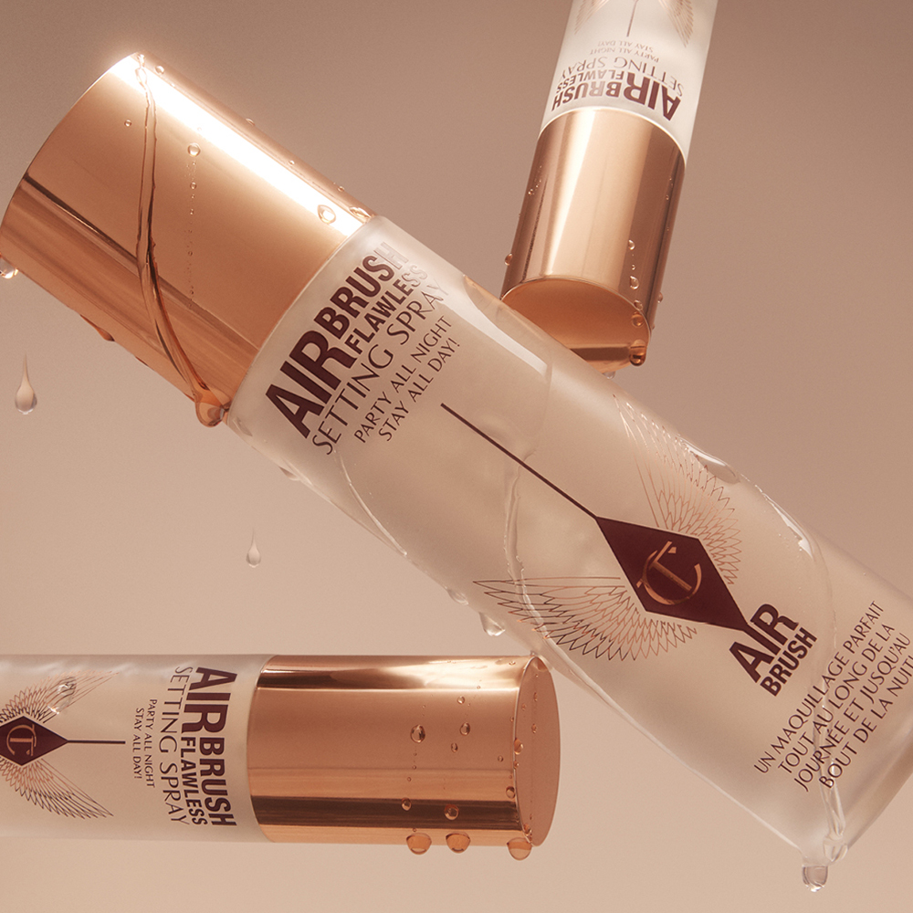 Three bottles of Airbrush Flawless Setting Spray with gold caps and water droplets on a beige background.