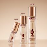 Three bottles of Airbrush Flawless Setting Spray in different sizes (34 ml, 100 ml, and 200 ml) with gold caps and a beige background.