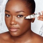 Deep-tone model with flawless, poreless, and glowy skin applying a white-coloured eye serum under her eyes that delivers a cooling sensation and moisturises.