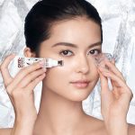 Fair-tone model with flawless, poreless, and glowy skin applying a white-coloured eye serum under her one eye and massaging an ice cube on the other side for a depuffing, cooling, and relaxing her skin.