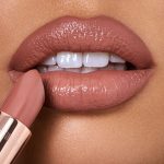 Lips close-up of a medium-tone model applying lipstick in a soft rosy peach shade with a satin finish.