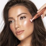 Medium-tone brunette model wearing glossy peach lipstick with shimmery gold and fawn eye makeup, and applying a glowy coral-peach blush from a liquid highlighter blush wand.