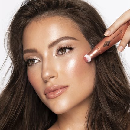 Medium-tone brunette model wearing glossy peach lipstick with shimmery gold and fawn eye makeup, and applying a glowy coral-peach blush from a liquid highlighter blush wand.