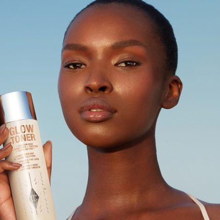 Deep-tone brunette model with glowy, luminous skin, having applied a glowy toner and holding up a bottle of it.