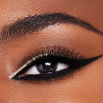 Single eye close-up of a deep-tone model with brown eyes wearing a duo eyeliner in black and champagne-beige on her upper lid and lower waterline.