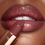 A dark, brownish-pink lipstick in front of a deep-skin model's lips that are coloured a glistening, brownish-pink.