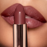 A deep, berry-pink lipstick in a metallic, golden tube held up in front of an olive skin model wearing the same shade.