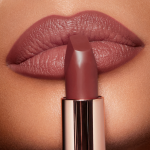 Close-up of a deep, berry-pink lipstick in a metallic golden tube, placed in front of the lips of a tanned model with lined lips wearing the same lipstick shade.