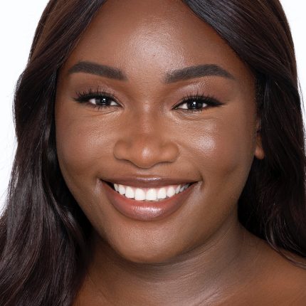 Deep-tone brunette model with brown eyes wearing nude pink lipstick with a skin-like foundation with a flawless, dewy finish.