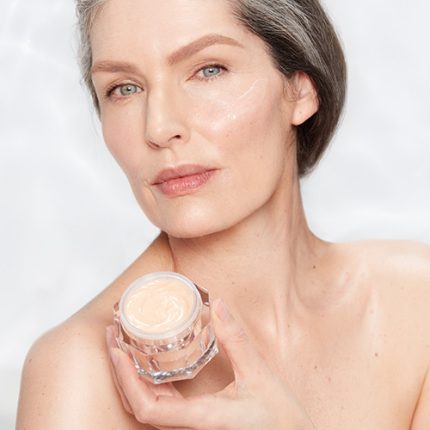 Fair-tone model with glowy, fresh, and mature skin, holding an open glass jar of a thick, peach-coloured night cream.