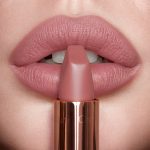 Lips close-up of a model wearing a matte, nude pink lipstick while holding the lipstick in front of her lips.