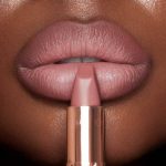 Lips close-up of a model wearing a matte, nude pink lipstick while holding the lipstick in front of her lips.