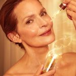 Fair-tone model with mature yet glowy, luminous, glass skin, applying a light-gold-coloured facial oil from a glass bottle with a gold and white-coloured dropper lid.