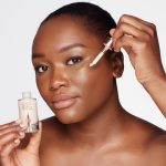 A deep-tone model with brown eyes wearing subtle eye makeup and applying a luminous face serum on her face with a gold-coloured dropper.