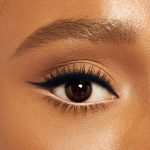 Close-up of a deep-tone model with brown eyes wearing jet-black eyeliner pencil on her eyelid and nude beige eyeliner pencil on her lower waterline.