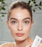 Light-tone model with hazel eyes and smooth, flawless, and glowy glass skin holding a white-coloured tube of a wash-off exfoliating mask.