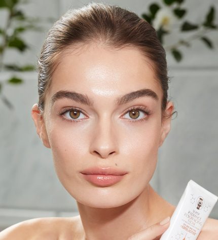 Light-tone model with hazel eyes and smooth, flawless, and glowy glass skin holding a white-coloured tube of a wash-off exfoliating mask.