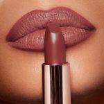 Lips close-up of a deep-tone model wearing a matte, berry-pink lipstick and holding the open lipstick in front of her lips.